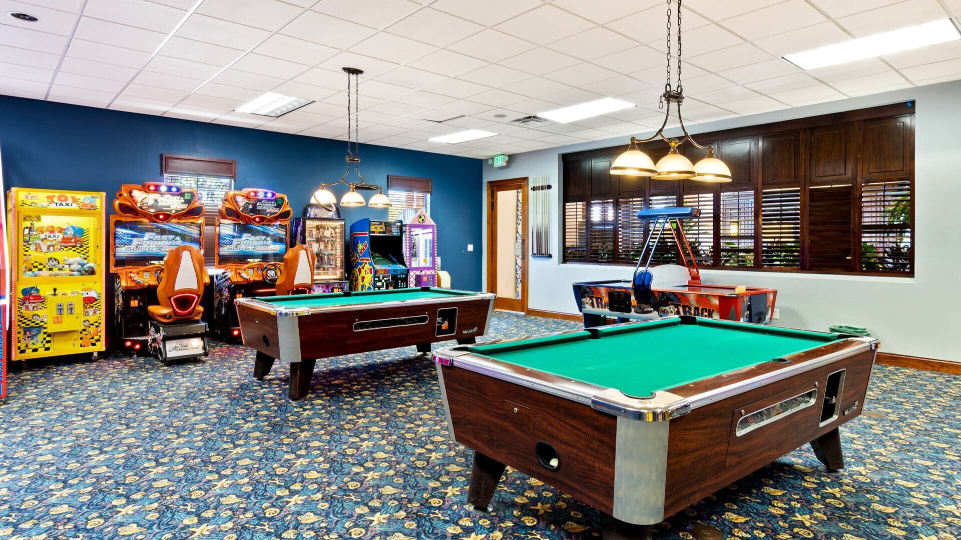 Une salle de billard avec deux tables de billard, des jeux d’arcade et une grande fenêtre équipée de stores.