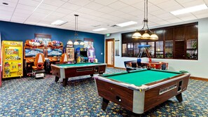 Un salón de billar con dos mesas de billar, juegos de arcade y un amplio ventanal con persianas.
