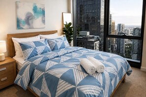 A hotel room with a large bed, a nightstand, a bedside lamp, a potted plant, and a city view through a large window. - Sea & Skyline Views | Pool, Gym & Sauna (Southbank)
