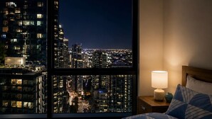 A hotel room with a large window offering a nighttime city view, a bed with patterned bedding, a bedside table with a lamp, and a nightstand. - Sea & Skyline Views | Pool, Gym & Sauna (Southbank)