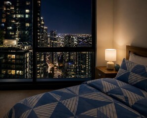 Ein Hotelzimmer mit großem Fenster, das einen nächtlichen Blick auf die Stadt bietet, einem Bett mit gemusterten Betttextilien, einem Nachttisch mit Lampe und einem weiteren Nachtisch.