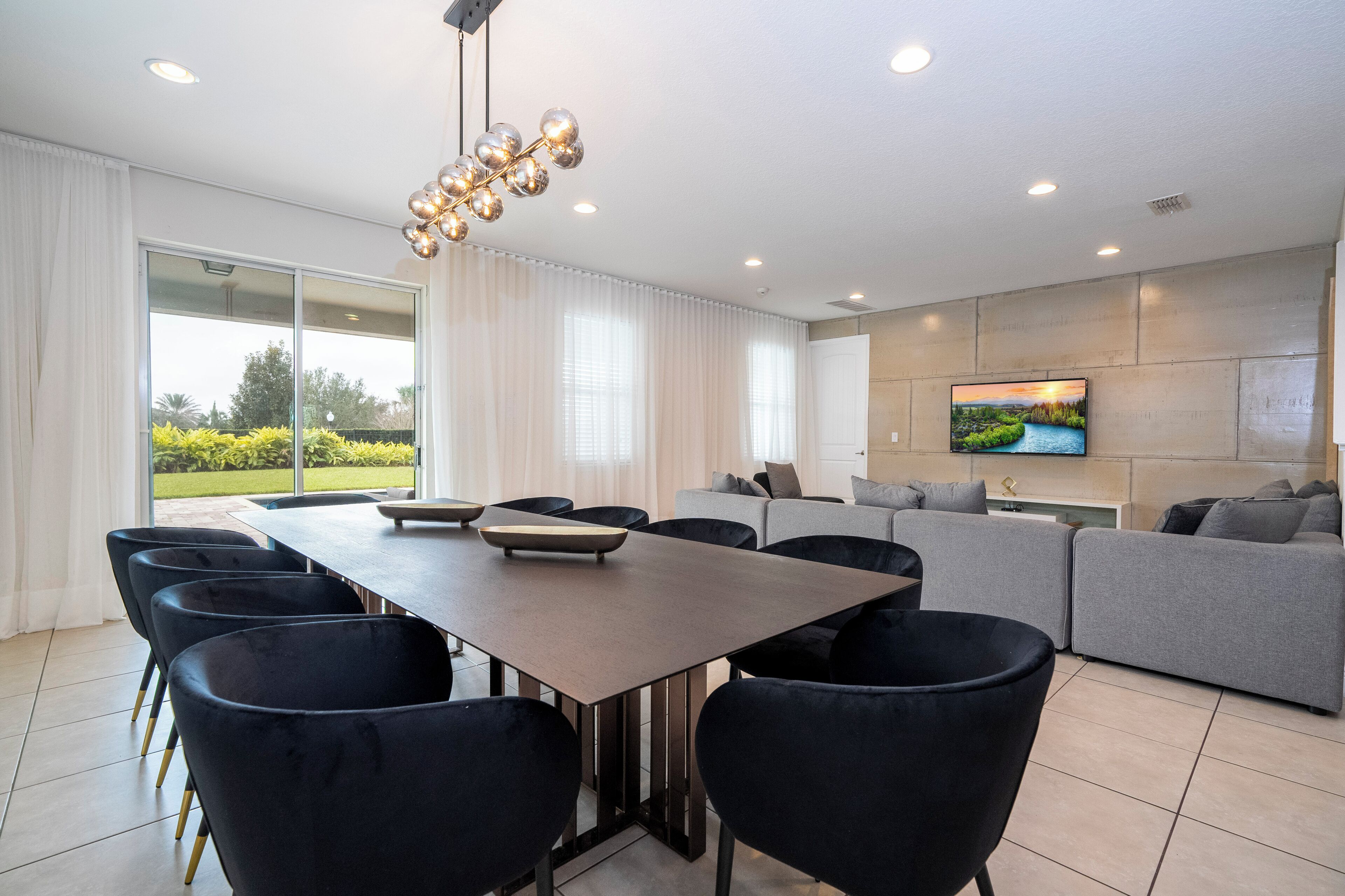 A modern living room with a dining area, a sofa set, a flat-screen TV mounted on the wall, and a view of the outdoors through large windows.
