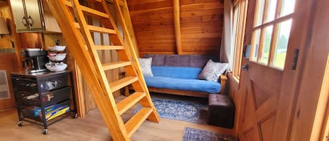 Un intérieur en bois avec un lit en mezzanine, un canapé, une échelle et une petite table avec des ustensiles de cuisine.