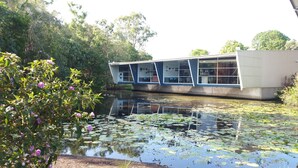 A building with a modern architectural design, situated beside a body of water with lily pads. - Villa 1 - Glass House Mountains Retreat (Beerwah)