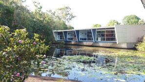 A building with a modern architectural design, situated beside a body of water with lily pads. - Villa 1 - Glass House Mountains Retreat (Beerwah)