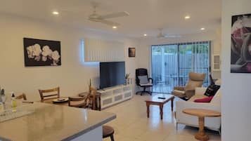 A spacious living area with a dining table, chairs, a sofa, a TV, and a balcony with sliding doors.