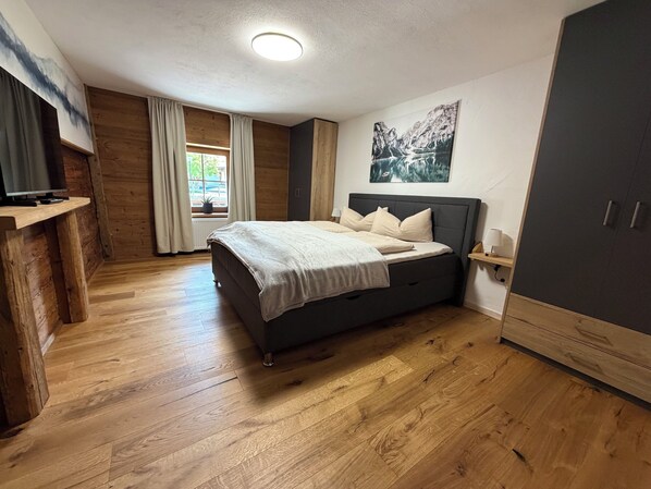 A bedroom with a bed, wooden floor, a desk, a window with curtains, and a painting on the wall.