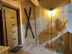 A wooden interior with a door, skis leaning against the wall, a lamp, and a vase with dried plants.