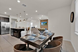 Une cuisine moderne dotée d’un îlot central, d’appareils électroménagers en acier inoxydable, de luminaires suspendus et d’un coin repas avec une table en bois prévue pour quatre personnes.
