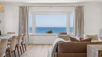 Een kamer met een groot raam dat uitzicht geeft op de zee, een eethoek met rieten stoelen, en een bank met kussens.