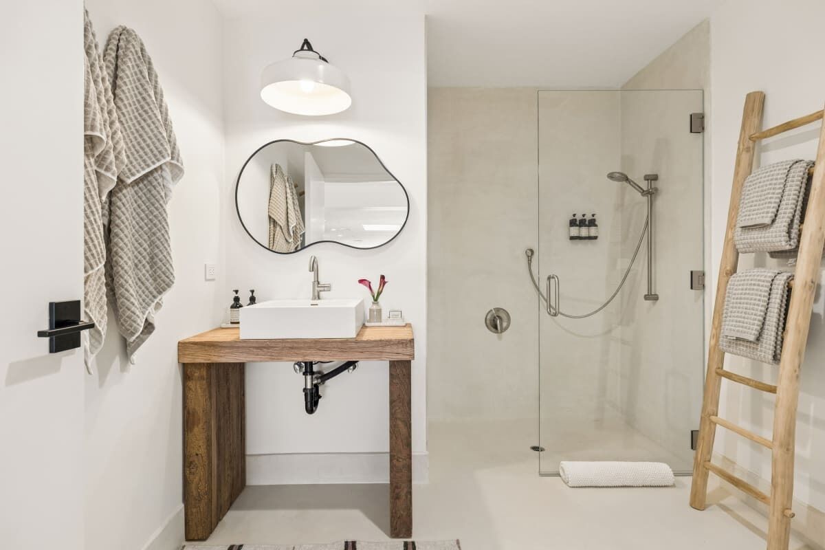 A modern bathroom with a wooden vanity, a round mirror, a glass shower enclosure, and a wooden ladder.