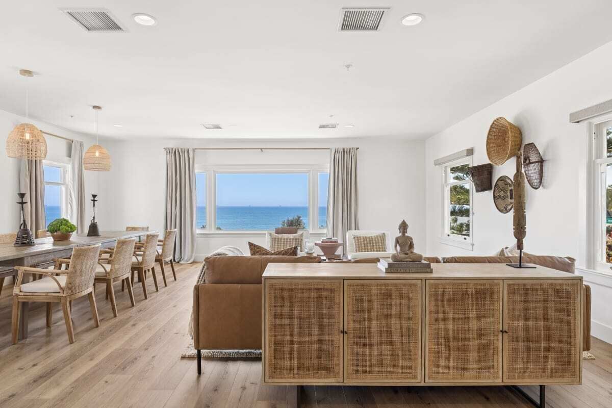 Een ruime kamer met een groot raam met uitzicht op zee, een eethoek met rieten meubels, en een centraal geplaatst rotan console.
