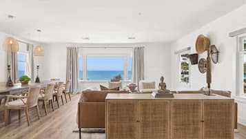 Een ruime kamer met een groot raam met uitzicht op zee, een eethoek met rieten meubels, en een centraal geplaatst rotan console.