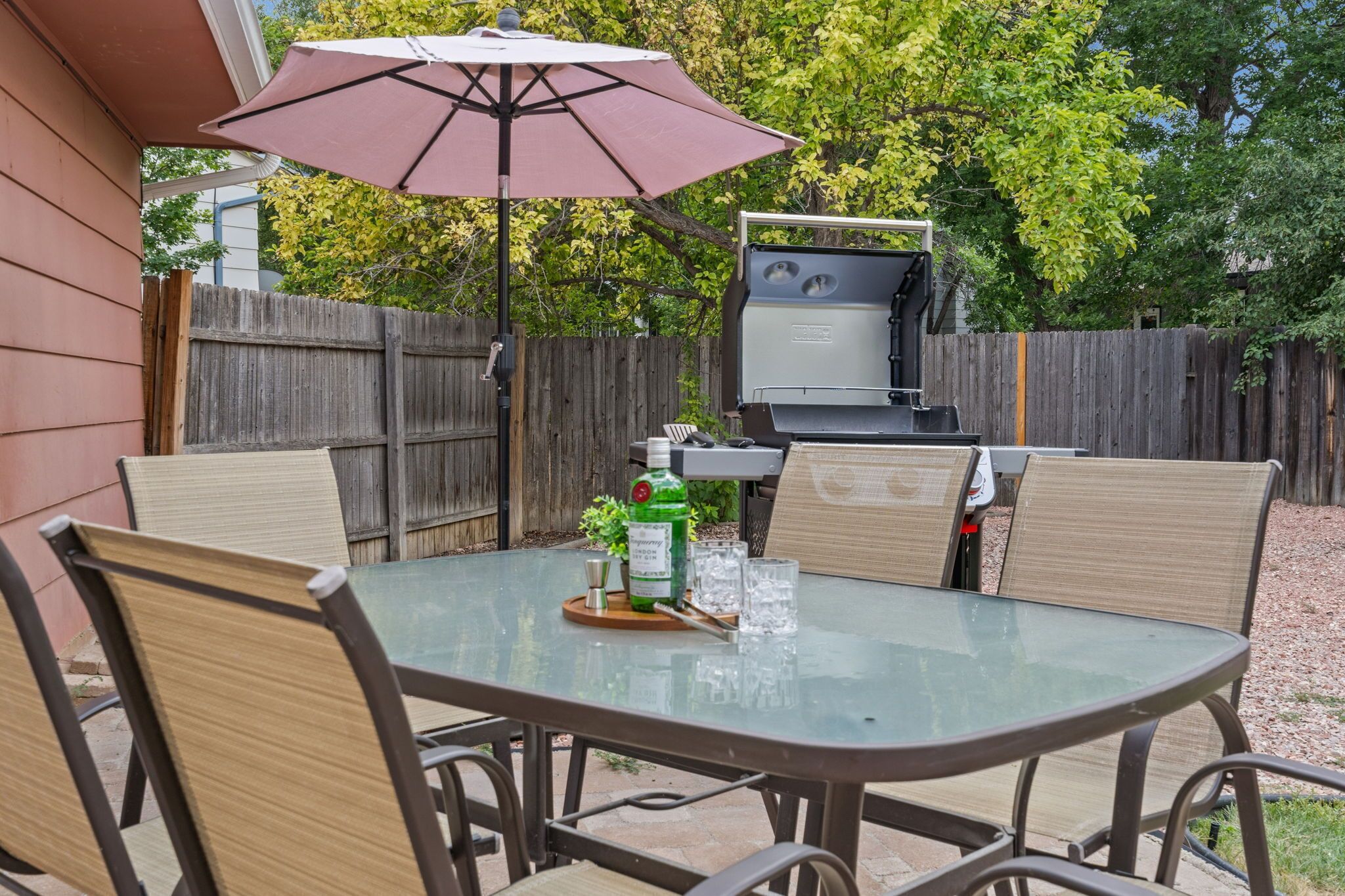 An outdoor patio with a glass table, four chairs, a barbecue grill, and a table umbrella.