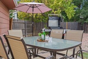 An outdoor patio with a glass table, four chairs, a barbecue grill, and a table umbrella. - Pipit Place - Pet-friendly Home w/ Fenced Yard (Fort Collins)