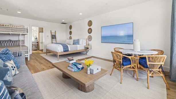 Una sala moderna con sofá, una mesa de centro, un comedor con sillas de mimbre, una cama y un amplio ventanal con vista al mar.