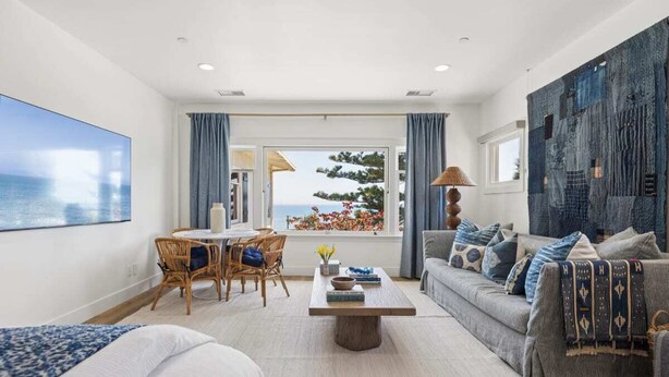 A modern living room with a view of the sea, a dining area with wicker furniture, and a cozy seating area with patterned cushions. - The Big Yellow House Loft B (Summerland)