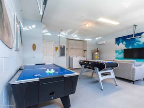 Ein Billardtisch, ein Kicker, eine Couch und ein an der Wand befestigter Fernseher in einem Raum mit weißen Wänden und einem gefliesten Boden.