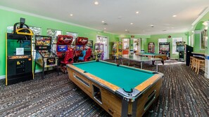 Una mesa de billar rodeada de juegos de arcade en una sala con paredes verdes y alfombra estampada.