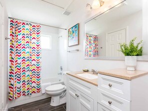 Un baño con inodoro blanco, cortina de ducha con estampado en zigzag, lavabo con encimera color beige y espejo sobre el lavabo.