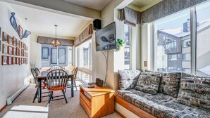 A modern living room with a sofa, a dining table with chairs, a TV mounted on the wall, and a bookshelf filled with books. - Ski-In/Ski-Out Kirkwood Village Condo (Kirkwood)