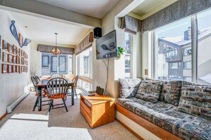 A modern living room with a sofa, a dining table with chairs, a TV mounted on the wall, and a bookshelf filled with books. - Ski-In/Ski-Out Kirkwood Village Condo (Kirkwood)