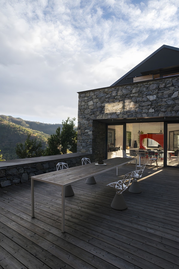 A modern outdoor dining area with a stone wall, wooden deck, and a view of the surrounding landscape.