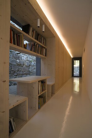 A modern interior with wooden shelving units filled with books, a stone wall, and a long hallway leading to an outdoor area.