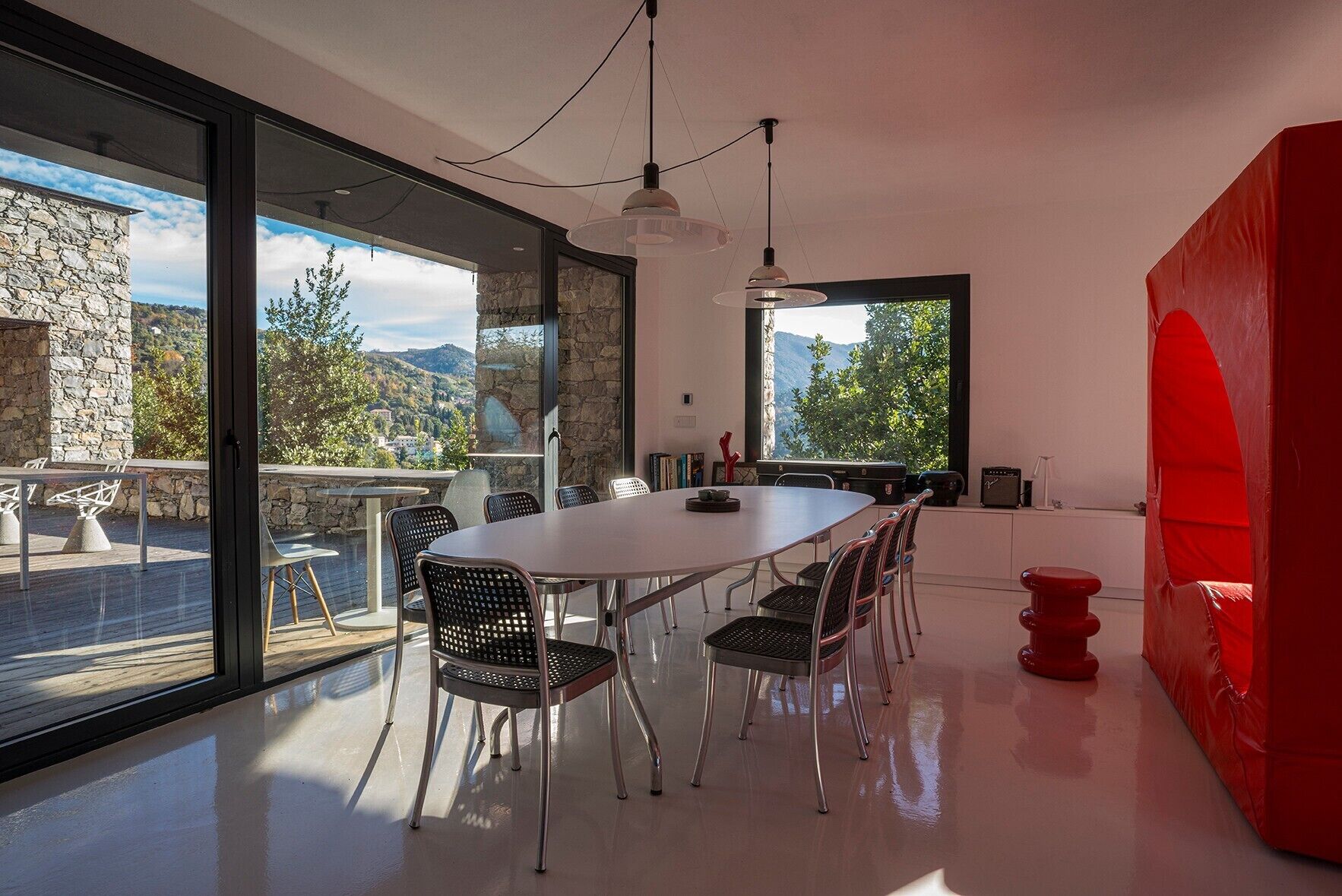 A modern dining area with a large table and chairs, a view of the outdoors, and a red accent wall.