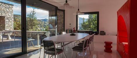 A modern dining area with a large table and chairs, a view of the outdoors, and a red accent wall.