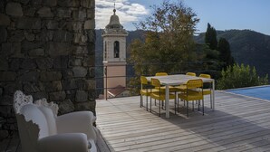 A wooden deck with a table and chairs, a stone wall, and a bell tower in the background.