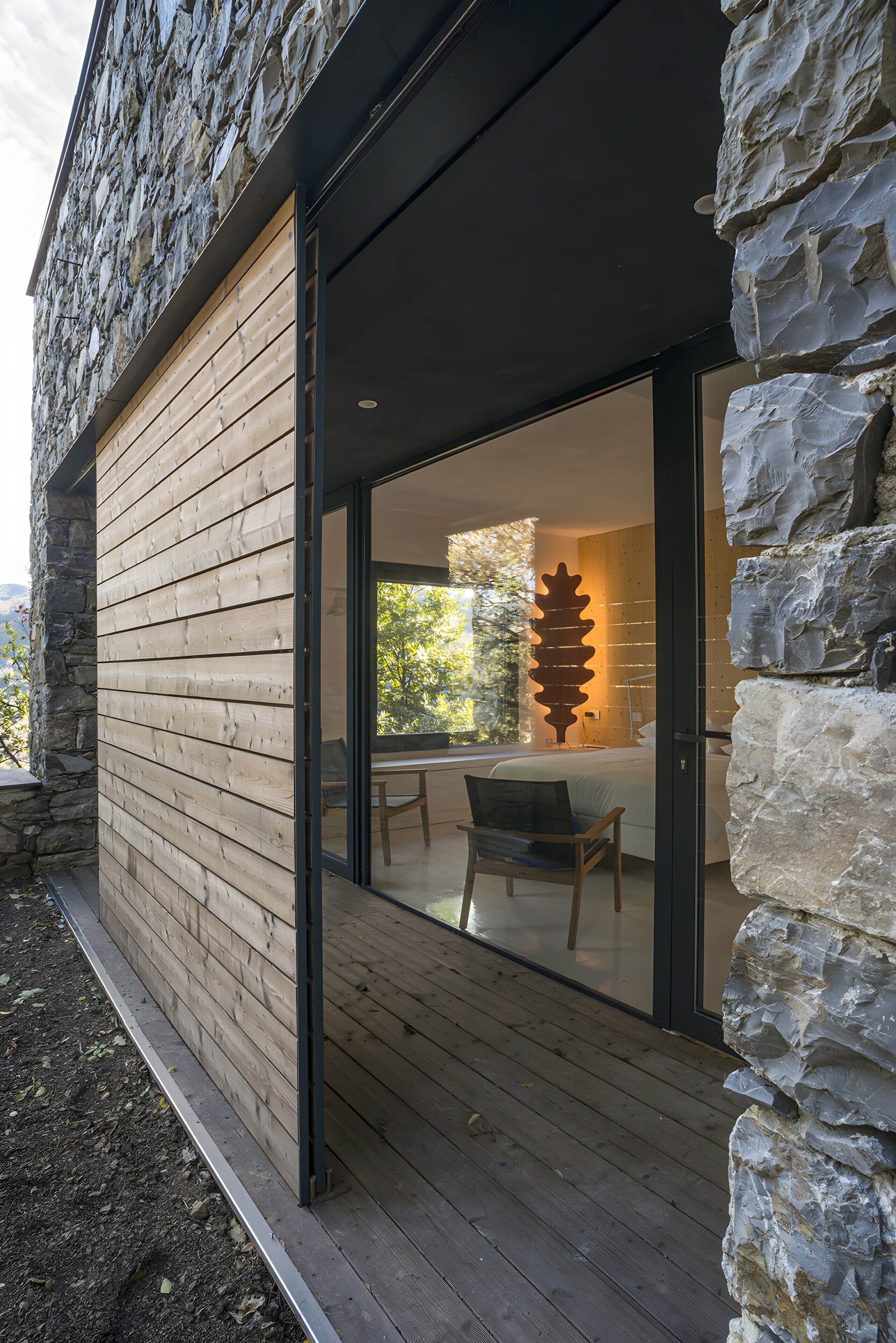 A modern building with a stone facade, wooden slats, and a glass door leading to an interior with a dining area and a large rock sculpture.