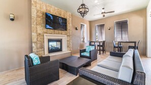 A modern living room with a stone wall, a fireplace, and a flat-screen TV mounted above it. There are wicker sofas and a dining area with a table and chairs. - Last Minute Disney! Private Pool, Hot Tub & Themed Bedrooms. Book Now! #107657 (Orlando)