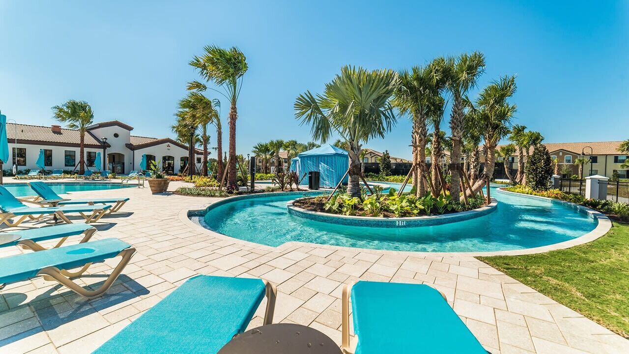 Un área de piscina con sillones de descanso, palmeras y una carpa azul.
