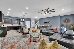 A modern kitchen with dark cabinets, a dining area with a table and chairs, a living room with a sofa and a coffee table, and a ceiling fan. - Last Minute Disney! Private Pool, Game Room & Themed Bedrooms. Book Now! #108401 (Orlando)