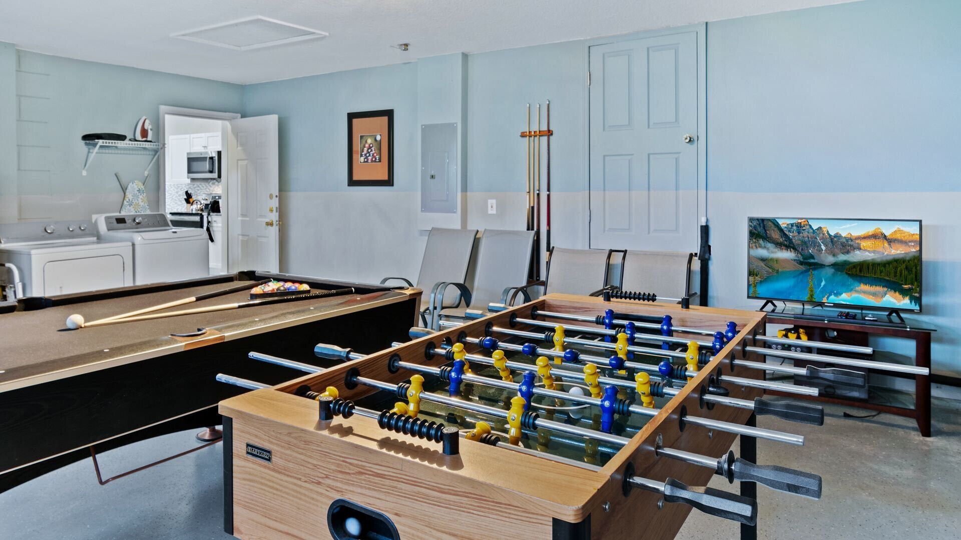 A room with a foosball table, a pool table, a television, and a laundry area.