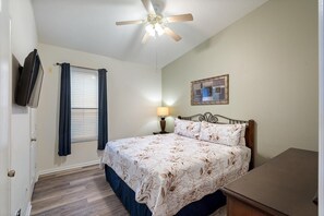 Quarto com cama, ventoinha no teto, janela com cortinas, abajur e uma obra de arte na parede.