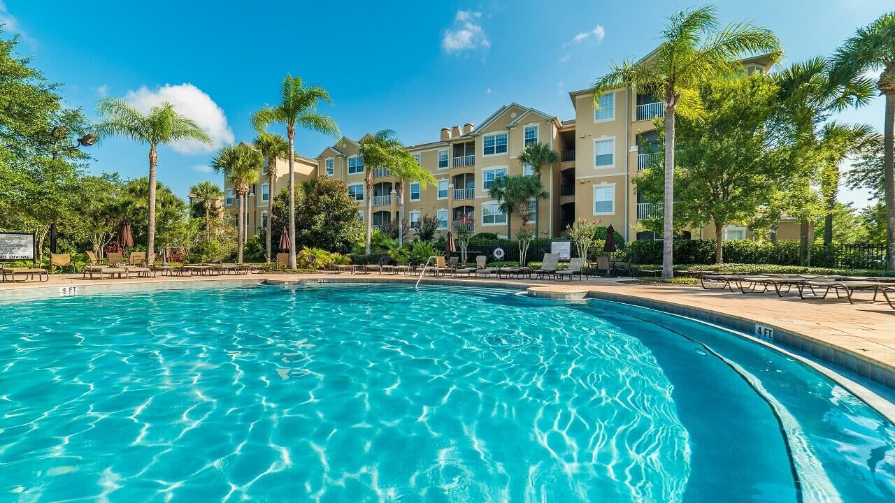 Une piscine à l’eau d’un bleu cristallin, entourée de palmiers et bordée d’un immeuble doté de nombreux balcons.