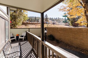 A covered outdoor area with a wooden deck, two white chairs, a table, and a view of a forested area with autumn foliage. - Premier Townhouse, Closest to the Slopes (Big Bear Lake)