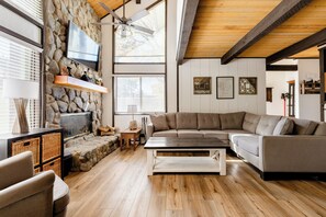 A modern living room with a stone wall, wooden ceiling beams, a large sectional sofa, and a flat-screen TV mounted on the wall. - Premier Townhouse, Closest to the Slopes (Big Bear Lake)