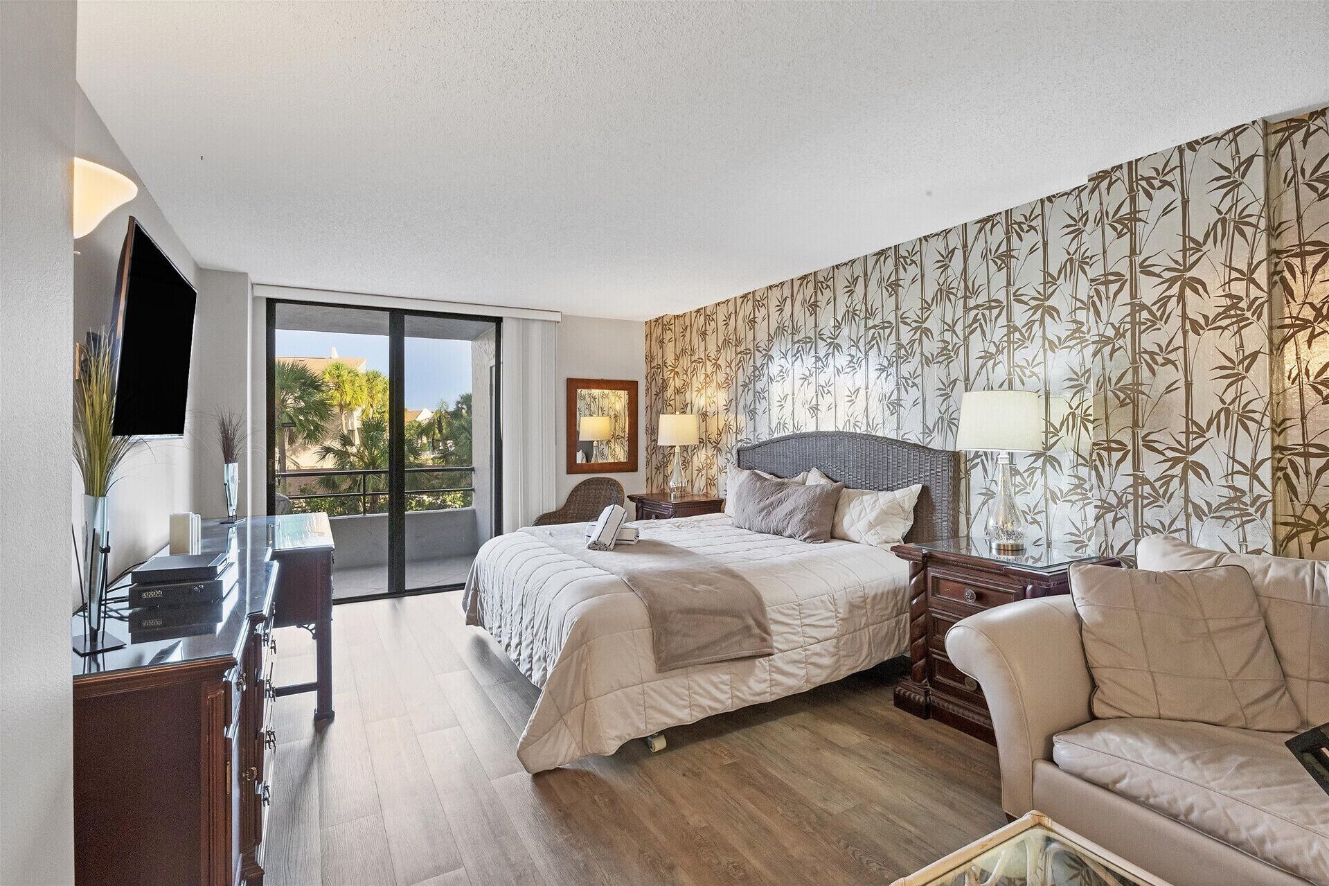 A hotel room with a bed, a sofa, a desk with a flat-screen TV, and a balcony with a view of palm trees.