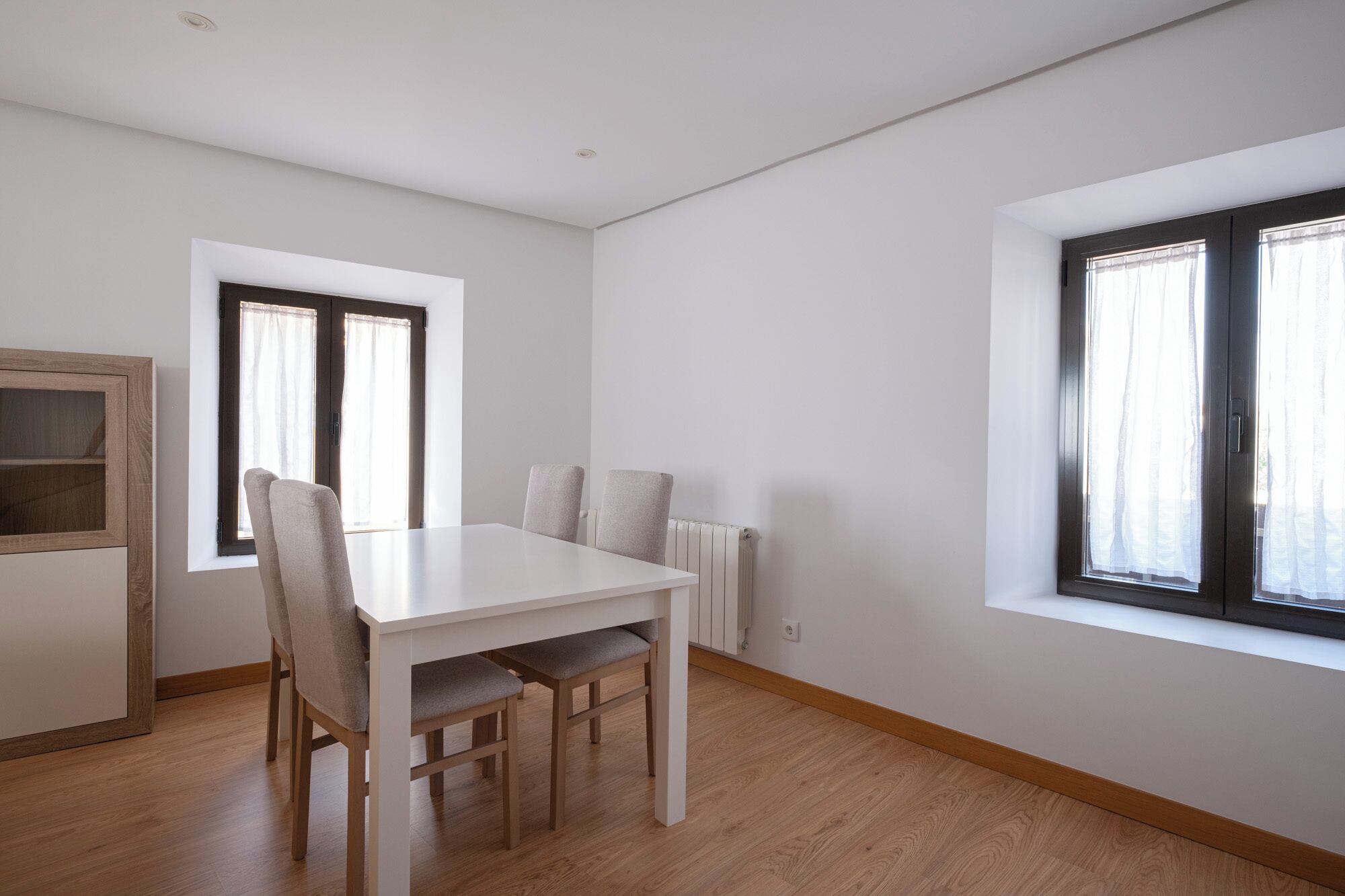 Ein Zimmer mit weißem Tisch, zwei Stühlen, einem Heizkörper und zwei mit Vorhängen versehenen Fenstern.