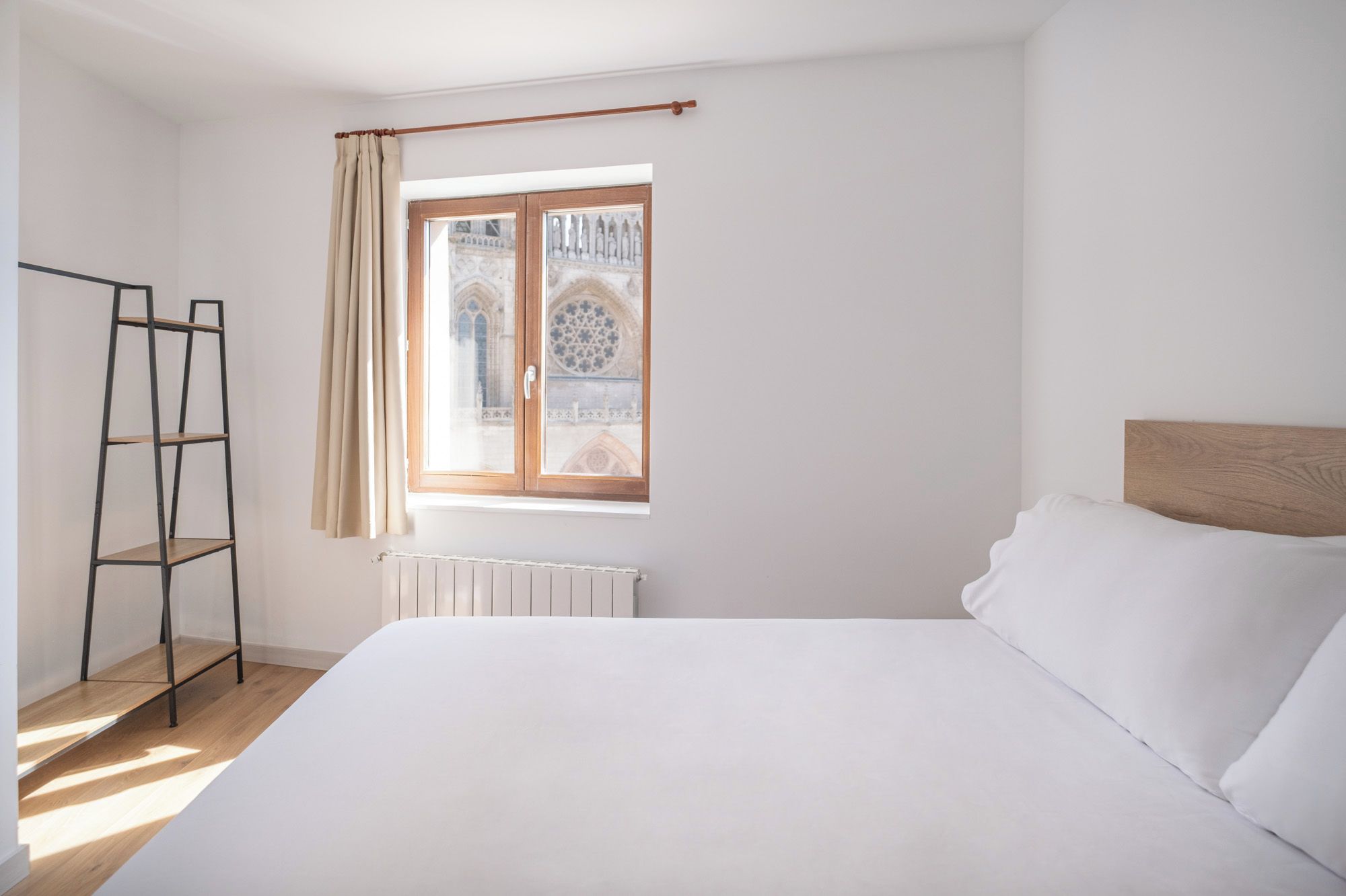 A bedroom with a bed, a radiator, a window with a view of a building, and a wooden ladder against the wall.