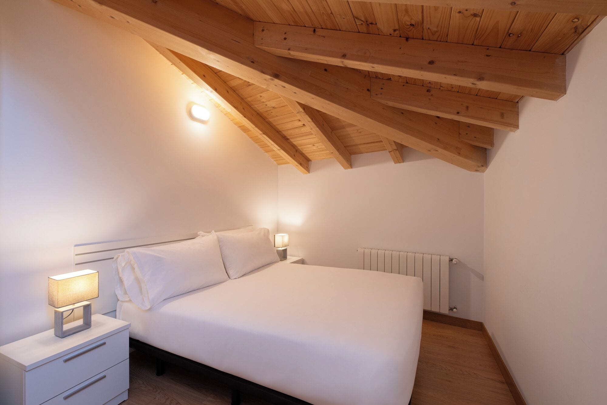 Una camera da letto con soffitto spiovente, un letto con biancheria bianca, un comodino con una lampada e un termosifone.