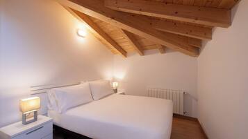 Una camera da letto con soffitto spiovente, un letto con biancheria bianca, un comodino con una lampada e un termosifone.