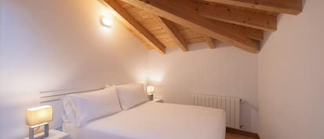 A bedroom with a sloped ceiling, a bed with white bedding, a bedside table with a lamp, and a radiator.