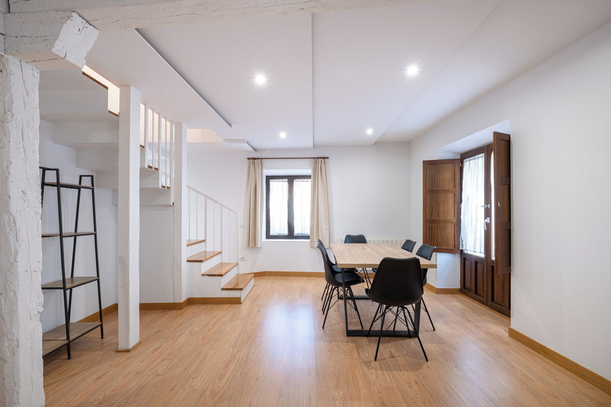 A spacious room with a wooden floor, a dining table with chairs, a staircase, and a large window with curtains.