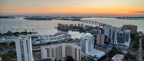 Un paisaje urbano costero con rascacielos, un puerto deportivo con barcos y un puente a lo lejos.
