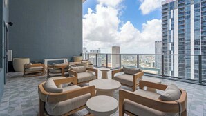 Uma área de estar moderna ao ar livre, com várias cadeiras e mesas, um vaso de planta grande e vista para a cidade.