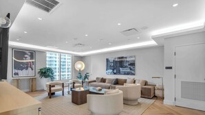 Uma sala de estar moderna com um conjunto de sofá, mesa de centro e poltronas. Há janelas amplas que proporcionam luz natural e vista para a cidade.